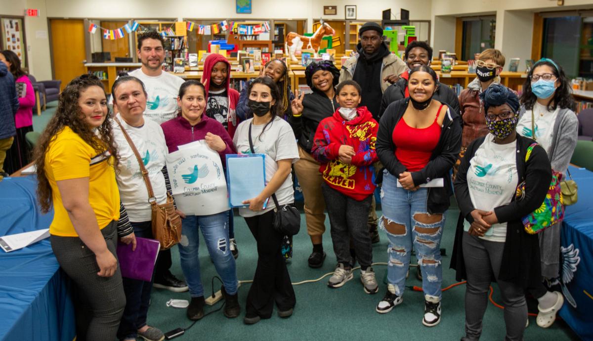 A crowd of people inside a library smiling at the camera and holding the CCSM logo